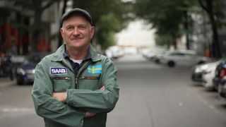 Autoexperte Andreas Keßler steht auf einer Straße mit einem grünen Overall und einer grauen Schiebermütze, blickt in die Kamera, die Arme verschränkt.