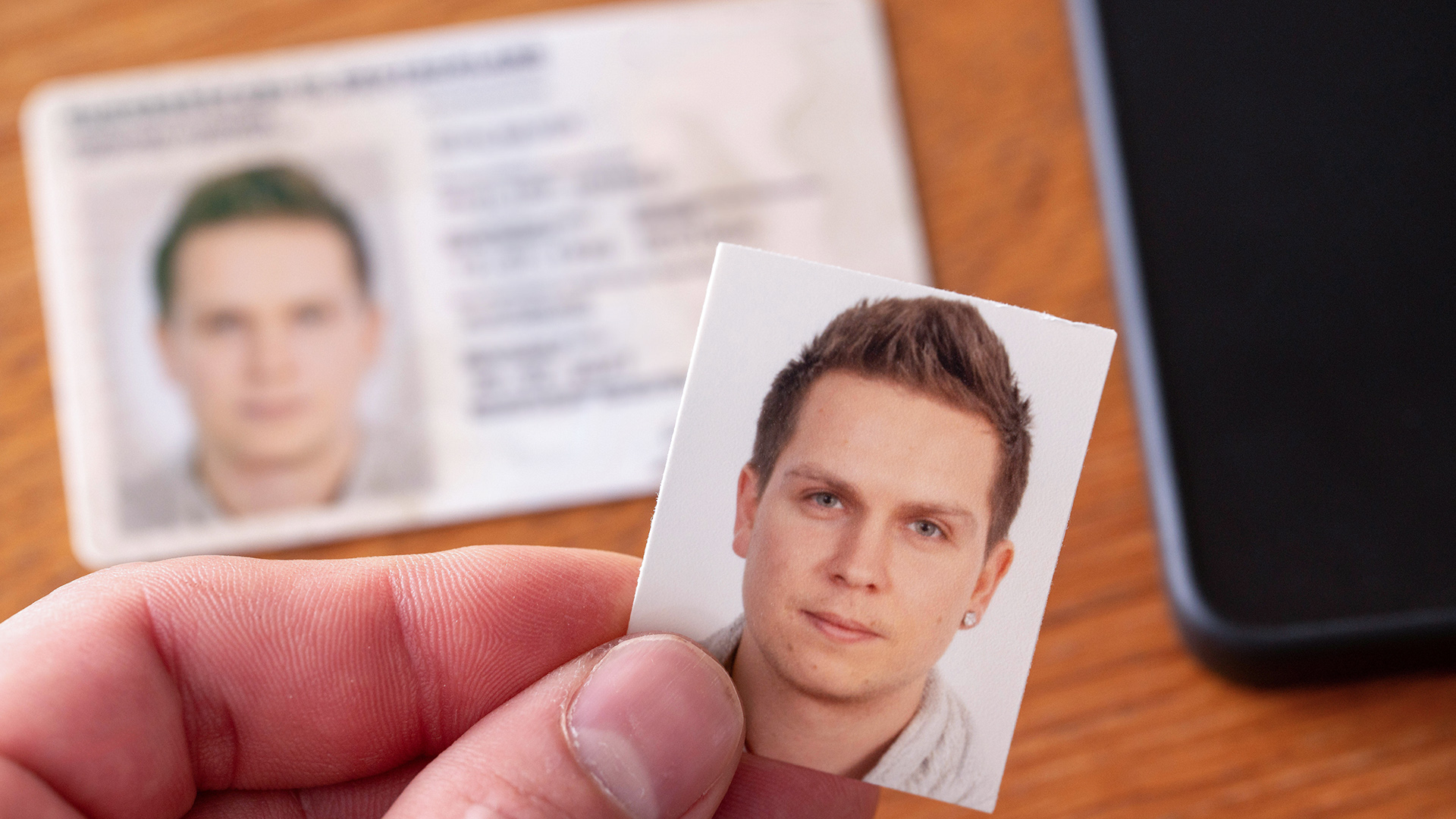 Symbolbild: Eine Hand hält ein Passbild vor einem Personalausweis und Handy auf Holztisch. (Quelle: dpa/Michael Bihlmayer)